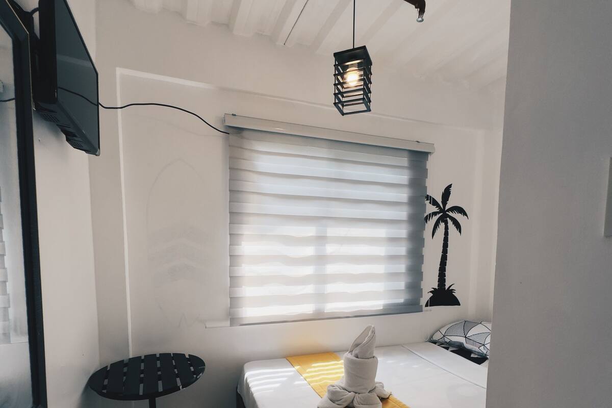 Une chambre d’hôtel compacte, comprenant un lit simple, une petite table, un téléviseur fixé au mur et une fenêtre avec des stores.