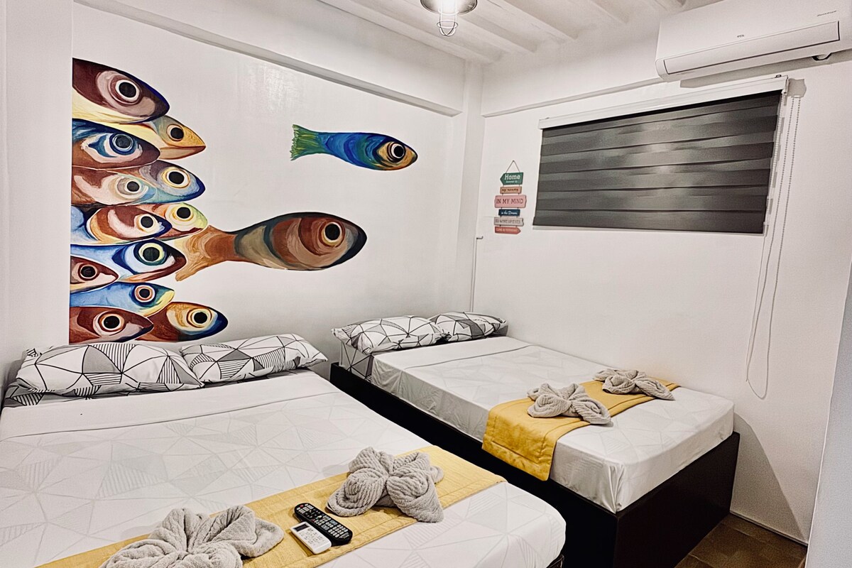 Ein Hotelzimmer mit zwei Betten, einem Deckenventilator, einem Bild von Fischen, einem Fenster mit Jalousien und einem kleinen Teppich.