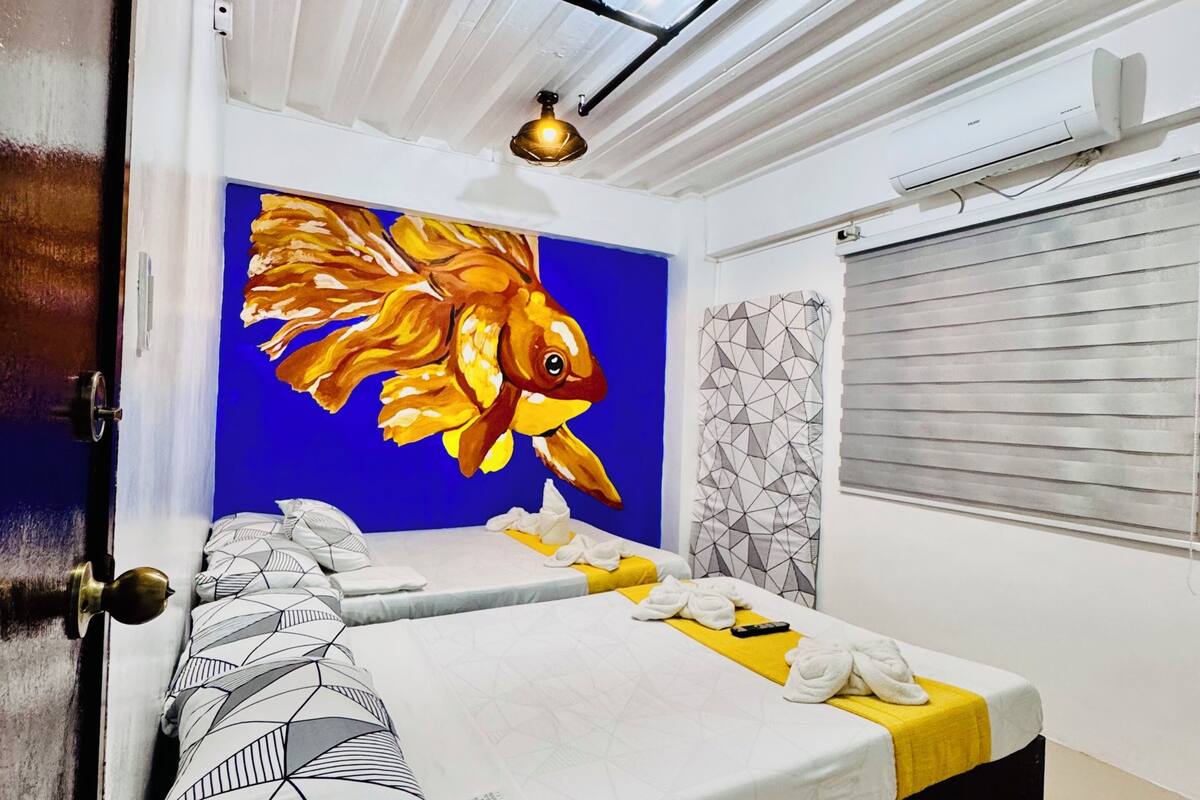 Une chambre d’hôtel avec un lit, un tableau représentant un poisson, un appareil de climatisation et une fenêtre avec des stores.