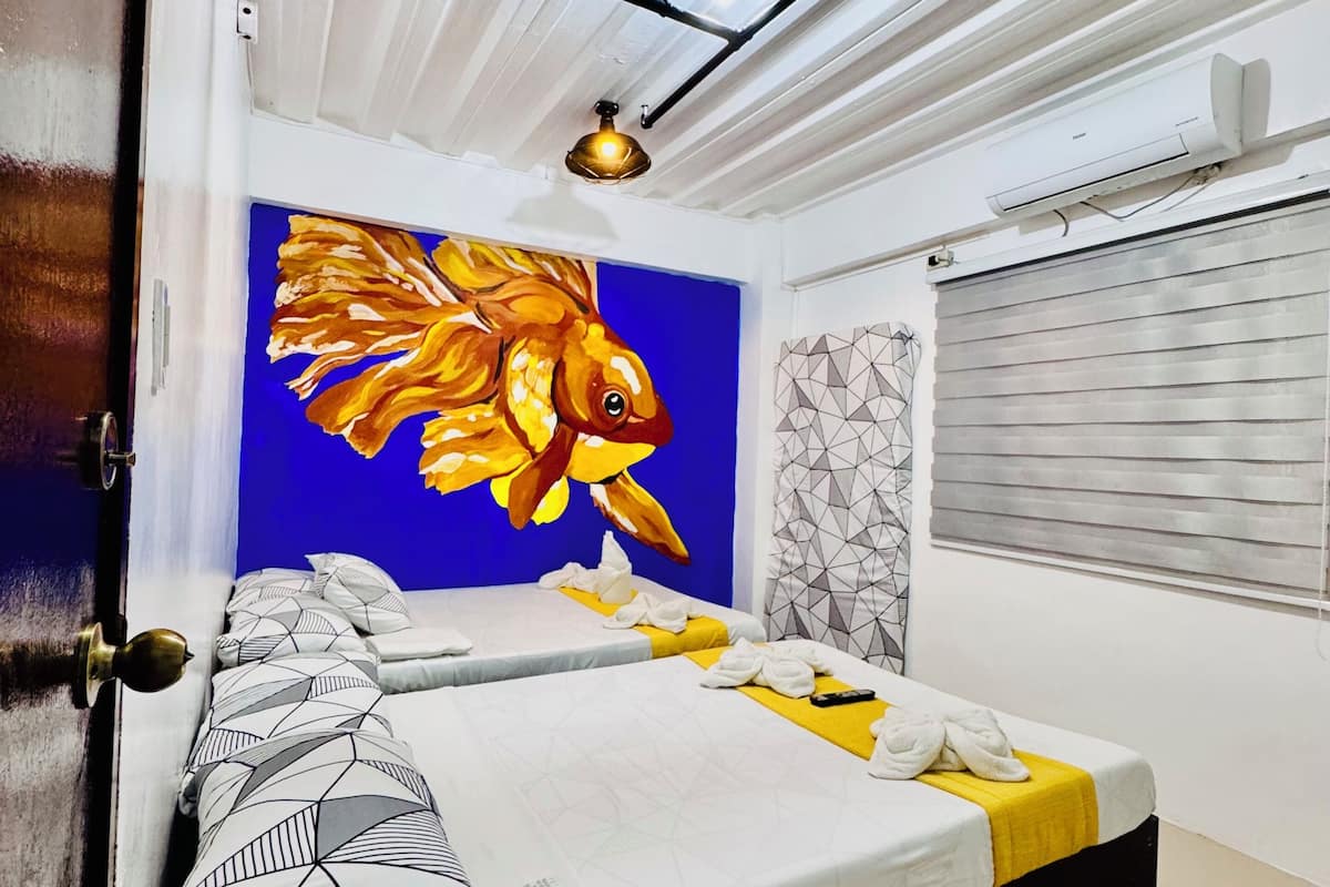 A hotel room with a bed, a painting of a fish, an air conditioning unit, and a window with blinds.