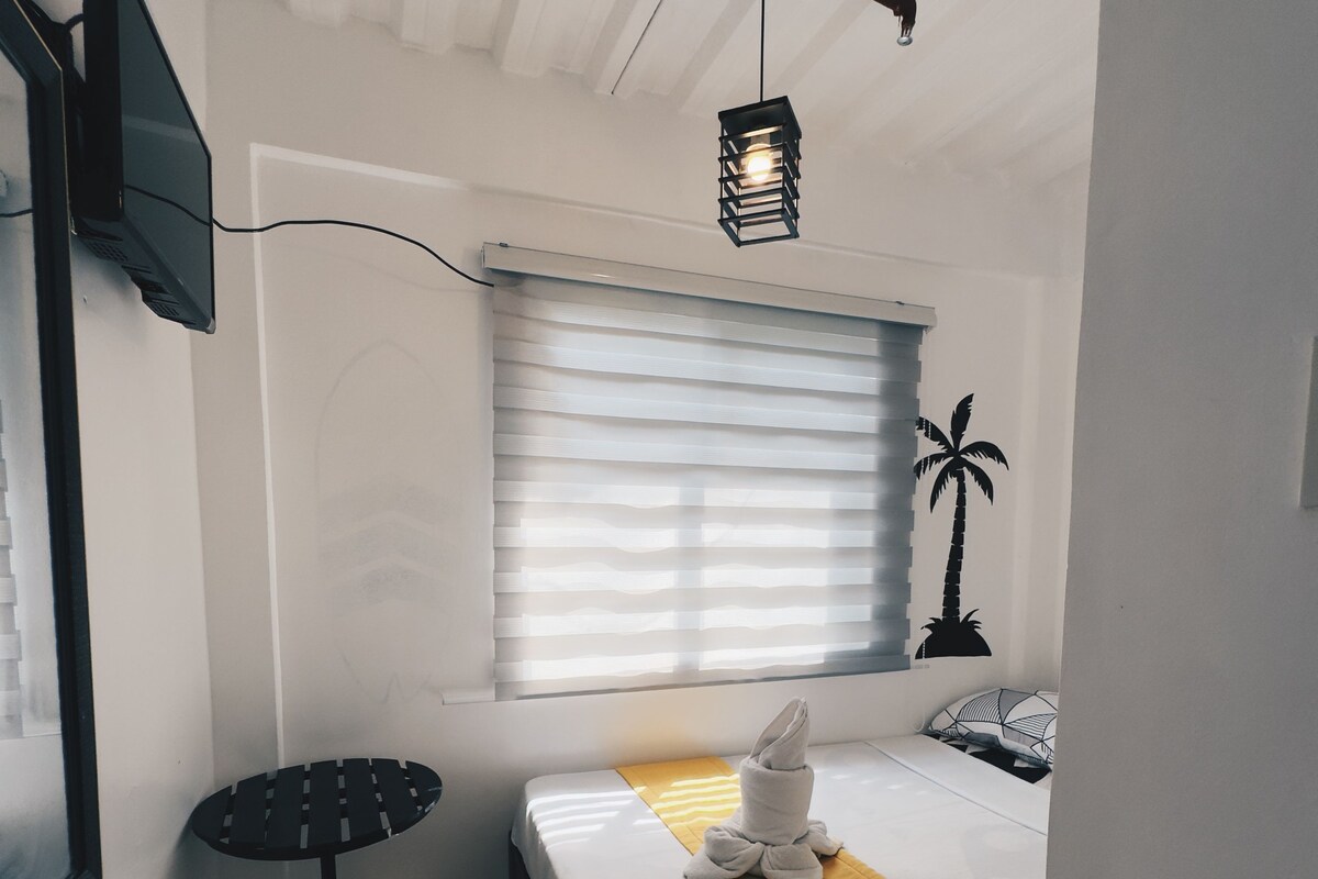 Ein kleines Hotelzimmer mit einem Einzelbett, einem kleinen Tisch, einem an der Wand befestigten Fernseher und einem Fenster mit Jalousien.
