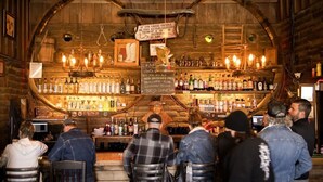 Um bar rústico com paredes de madeira, um arco de pedra e diversas garrafas e copos em prateleiras.