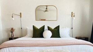 Quarto com cama, almofadas, manta fofa, espelho e luminárias na parede.
