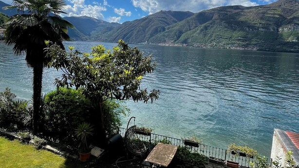 A lakeside patio with lounge chairs, a table, and a grill area, overlooking a calm lake and distant mountains. - Romantic studio with terrace overlooking the lake. (Lezzeno)