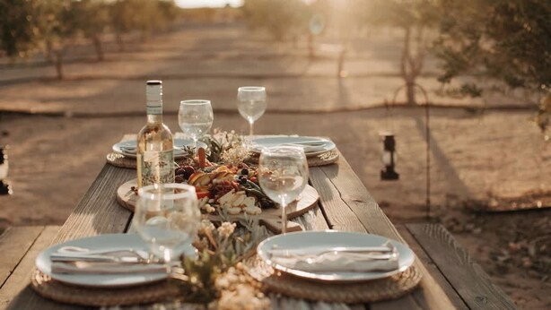 A wooden table set for a meal outdoors with plates, cutlery, and wine glasses.