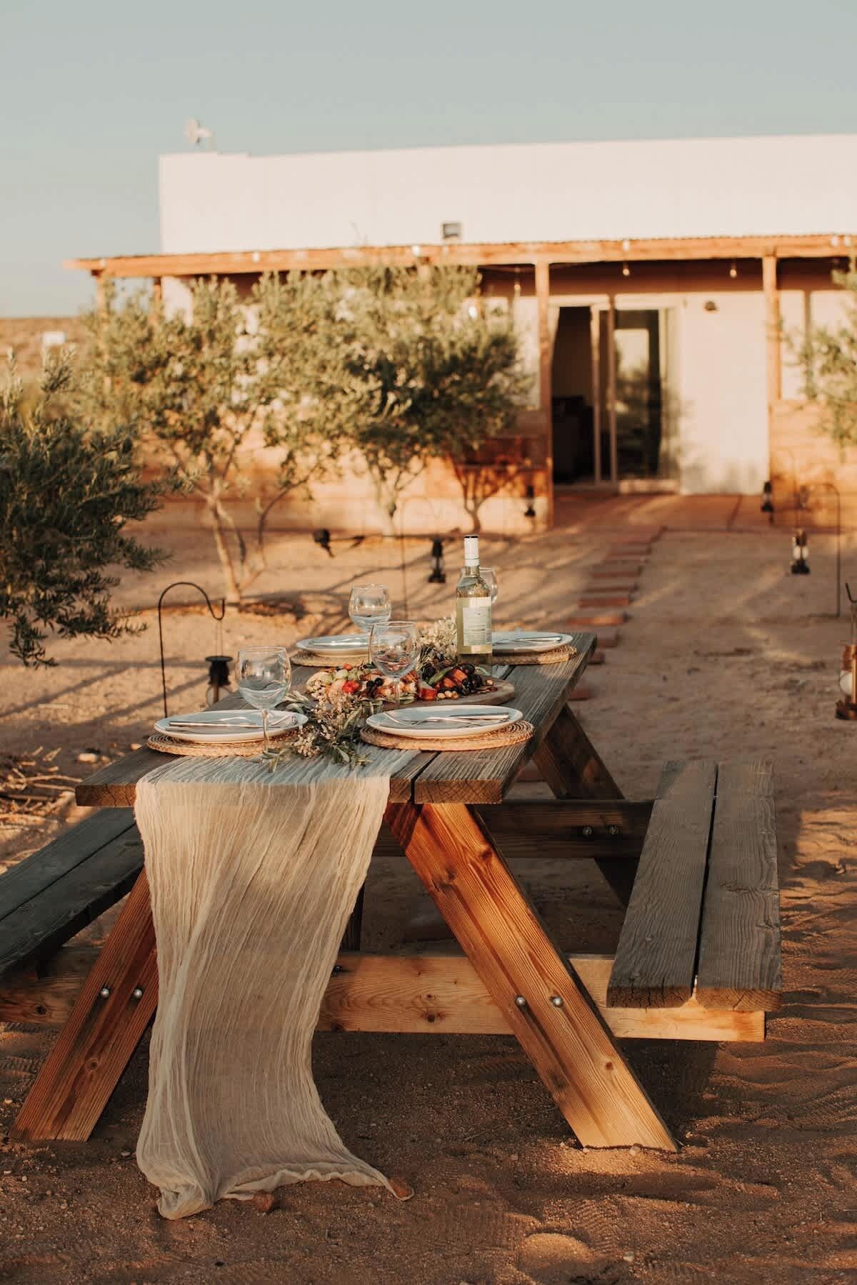 A rustic outdoor dining setup with a wooden table and benches, set against a building with a modern design and a clear sky.