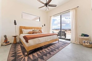 A bedroom with a bed, a ceiling fan, a large window with a view, and a sliding door leading to an outdoor area. - Noetic House · Hot Tub, Cowboy Tub, Views, 5 Acres (Joshua Tree)