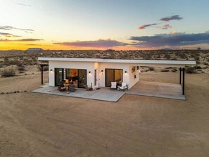 A modern, single-story house with a flat roof, large glass doors, and a patio area with outdoor furniture. - Noetic House · Hot Tub, Cowboy Tub, Views, 5 Acres (Joshua Tree)