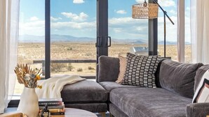 Ein modernes Wohnzimmer mit einem grauen Ecksofa, einem Holztisch und Blick auf eine Wüstenlandschaft durch große Fenster.