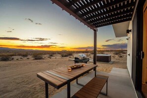 A covered outdoor patio with a wooden table and bench, a grill, and a view of a desert landscape at sunset. - Noetic House · Hot Tub, Cowboy Tub, Views, 5 Acres (Joshua Tree)