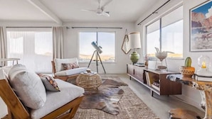 A spacious living room with a large window, a ceiling fan, a sofa, a coffee table, and a rustic rug. - Overlook JT • Pool & Spa, Sweeping Views Near Park (Joshua Tree)