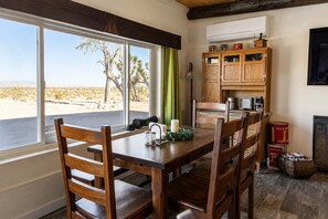 A dining area with a wooden table and chairs, a window with a view of a desert landscape, and a wooden cabinet with kitchen items. - Pipes Canyon Homestead • Privacy, Epic Views, Spa (Yucca Valley)