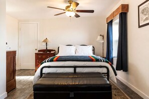 A bedroom with a bed, a bench, a ceiling fan, a nightstand, and a window with curtains. - Pipes Canyon Homestead • Privacy, Epic Views, Spa (Yucca Valley)
