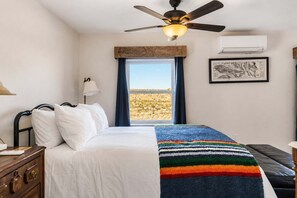 Ett hotellrum med en säng, en takfläkt, en luftkonditioneringsanläggning, ett fönster med utsikt över ett ökenlandskap och en inramad tavla på väggen.