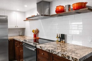 A kitchen with white tiled walls, a stainless steel refrigerator, wooden cabinets, and a granite countertop. - Pipes Canyon Homestead · Hot Tub, Views, 5 Acres (Yucca Valley)