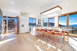 A modern kitchen with a large island, bar stools, and large windows offering a view of the mountains. - Stargate · 5 Acres, Pool, Hot Tub, Mountain Views (Yucca Valley)