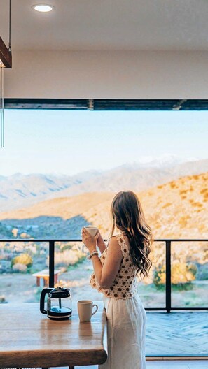 A person sitting at a table with a cup, overlooking a mountainous landscape through large windows. - Stargate · 5 Acres, Pool, Hot Tub, Mountain Views (Yucca Valley)