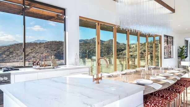 A modern kitchen with a marble island, large windows, and a view of mountains.