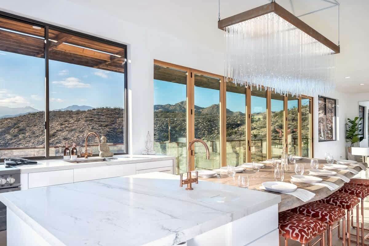 A modern kitchen with a marble countertop, a long dining table, and a view of a mountainous landscape through large windows.