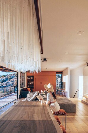 A modern interior with a large wooden table, a person reading, and a view of a mountainous landscape through large windows. - Stargate · 5 Acres, Pool, Hot Tub, Mountain Views (Yucca Valley)