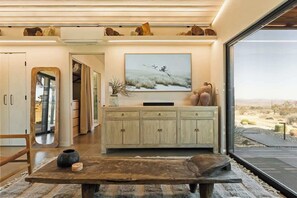 A modern living room with a large window, a wooden table, a cabinet with a flat-screen TV, and a painting of a landscape. - Shadow House • Serene Escape, Views, 10-Acres, Spa (Joshua Tree)