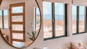 Una habitación con un mueble de diseño en patrón de espiga, un espejo redondo, un tocadiscos, un sofá con cojines, y vistas a un paisaje desértico a través de amplias ventanas.