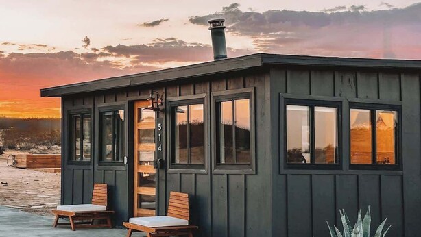Una cabaña de madera pequeña y oscura con dos sillas de madera y una mesa afuera, una chimenea y vistas del atardecer.