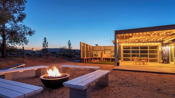 An outdoor patio area with a fire pit, benches, and a building with string lights. - Starship Landing • 2 Epic Houses Retreat, 10 Acres (Yucca Valley)