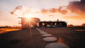 A modern building with a sunlit sky, a paved pathway leading to the entrance, and a desert landscape. - Starship Landing • 2 Epic Houses Retreat, 10 Acres (Yucca Valley)