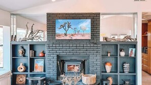 A modern living room with a fireplace, built-in shelving with decorative items, and a large window. - Stone&Sky • Pool & Spa in Boulders (Yucca Valley)