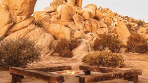 A circular fire pit with benches in front, surrounded by dry vegetation and large boulders. - Stone&Sky • Pool & Spa in Boulders (Yucca Valley)