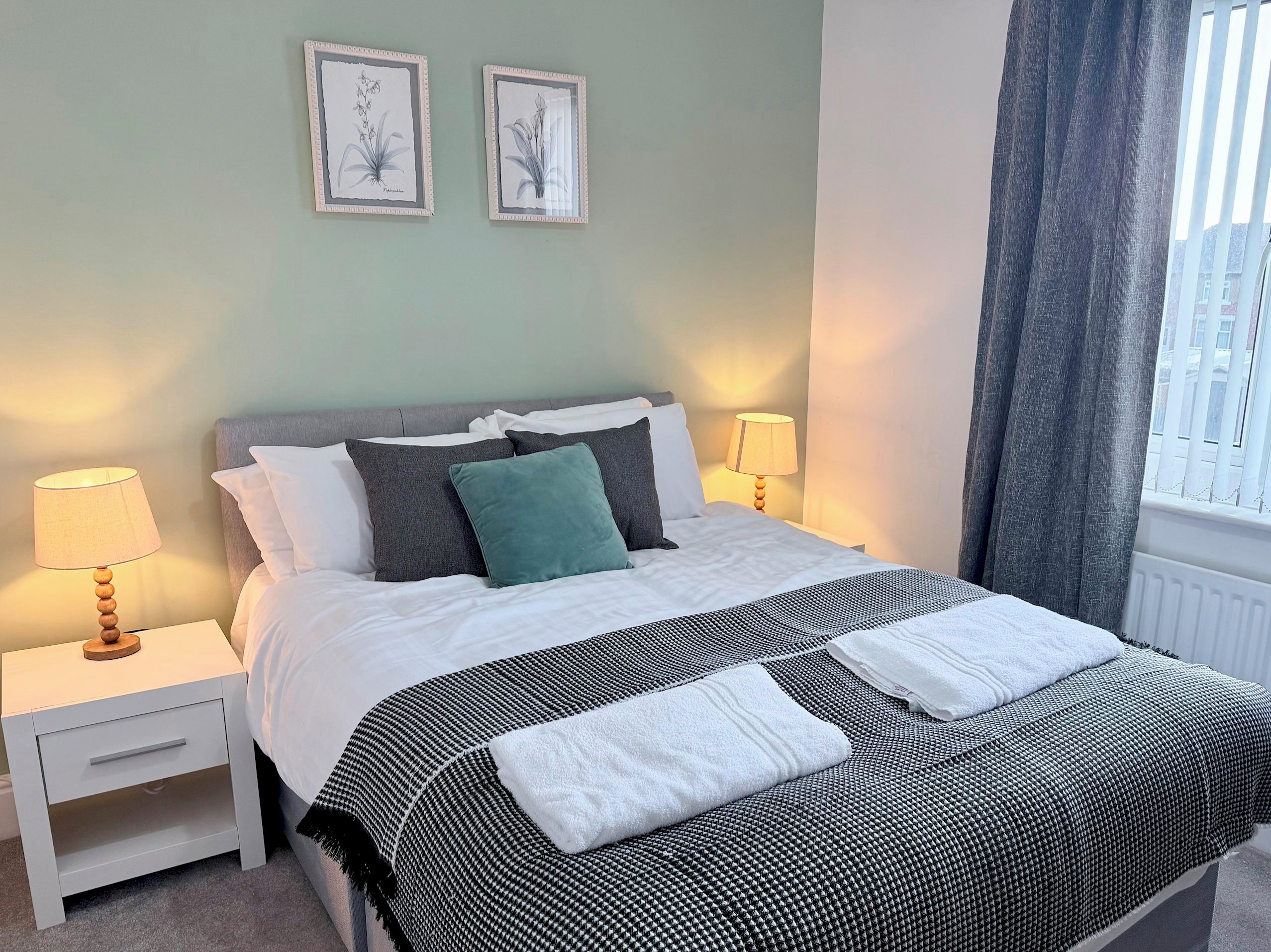 A neatly made bed with a black and white checkered bedspread, white pillows, and a teal accent pillow. There are two bedside tables with lamps, and two framed botanical prints on the wall.