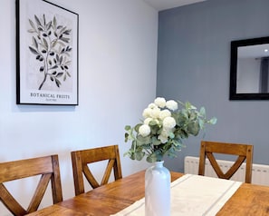 A dining table with a white vase of flowers, wooden chairs, a framed botanical print, and a mirror on the wall. - Toast Stays - The Stray Space (Darlington)