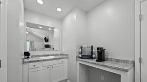 A modern bathroom with white cabinetry, a granite countertop, a sink, a mirror, a coffee maker, and a shower area.