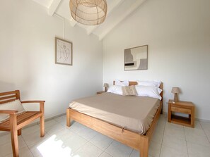 A bedroom with a wooden bed, a chair, a bedside table, and a hanging light fixture. - Villa Caméo • Grande villa familiale vue mer 180° (Le Gosier)