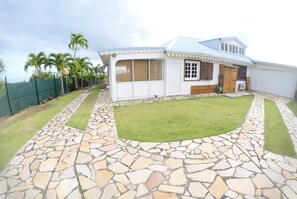 Une maison avec une terrasse en pierre, une pelouse et un espace clos agrémenté de palmiers.