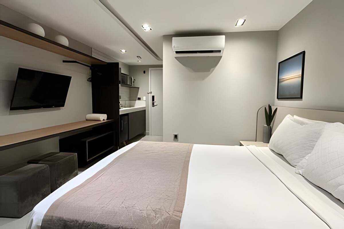 A modern hotel room with a bed, a TV, a kitchenette, and a plant.
