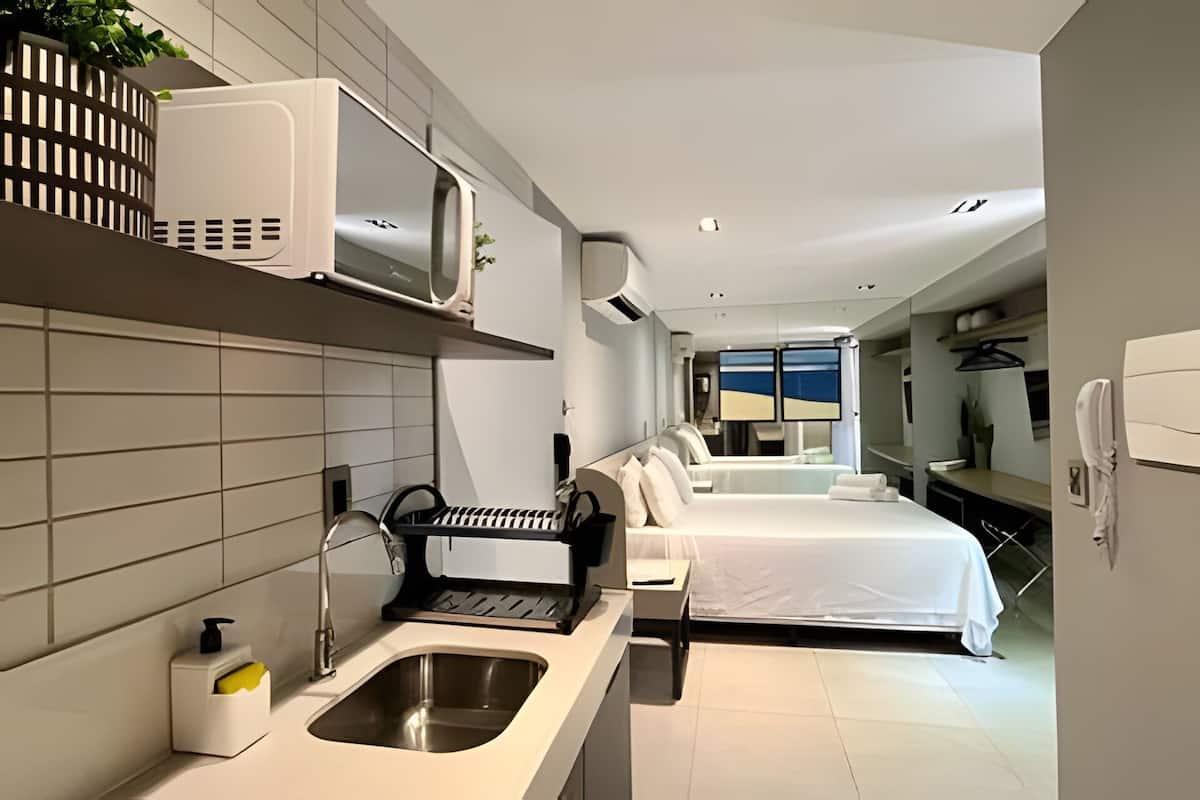 A modern hotel room with a kitchen area, a bed, a small desk, and a view of the sea.