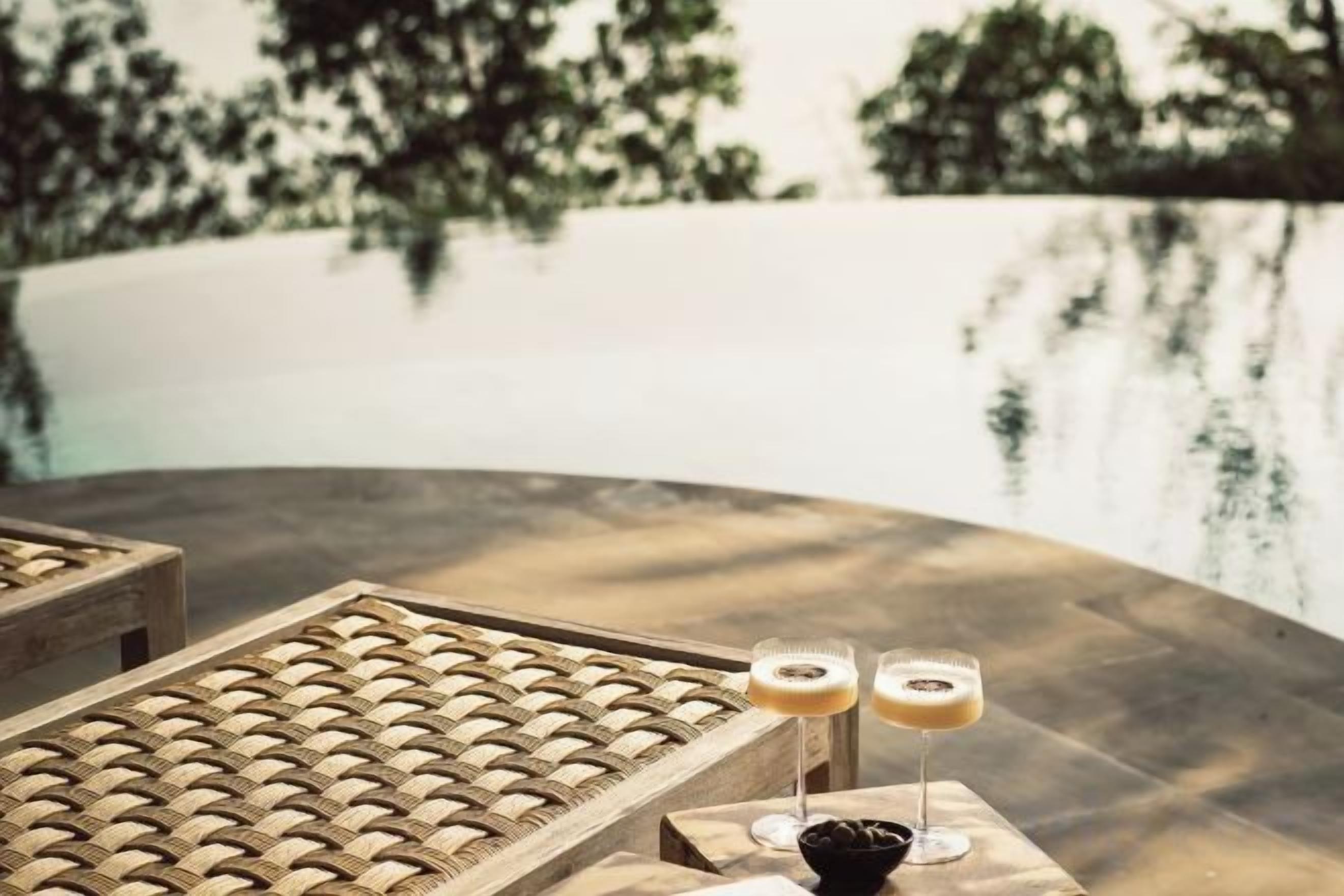 A wooden table with two glasses of a beverage, a wicker chair, and a view of a body of water with trees in the background.