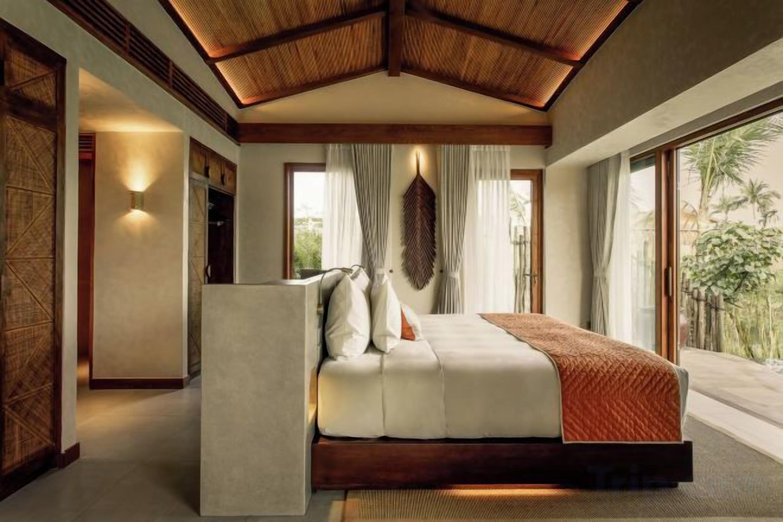 A bedroom with a large bed, a wooden ceiling, and a view of greenery outside.