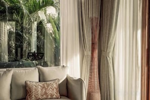 A room with a sofa, a window with curtains, and a view of palm trees. - Namia River Retreat - Wellness Inclusive Resort (Hoi An)
