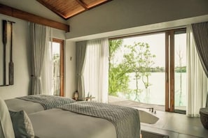 A bedroom with a large bed, a window with curtains, and a view of greenery. - Namia River Retreat - Wellness Inclusive Resort (Hoi An)