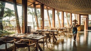 An open dining area with wooden tables and chairs, large windows, and a thatched ceiling. - Namia River Retreat - Wellness Inclusive Resort (Hoi An)