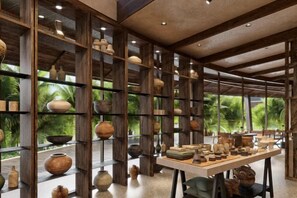 A room with shelves displaying pottery, a table with books, and a view of greenery outside. - Namia River Retreat - Wellness Inclusive Resort (Hoi An)