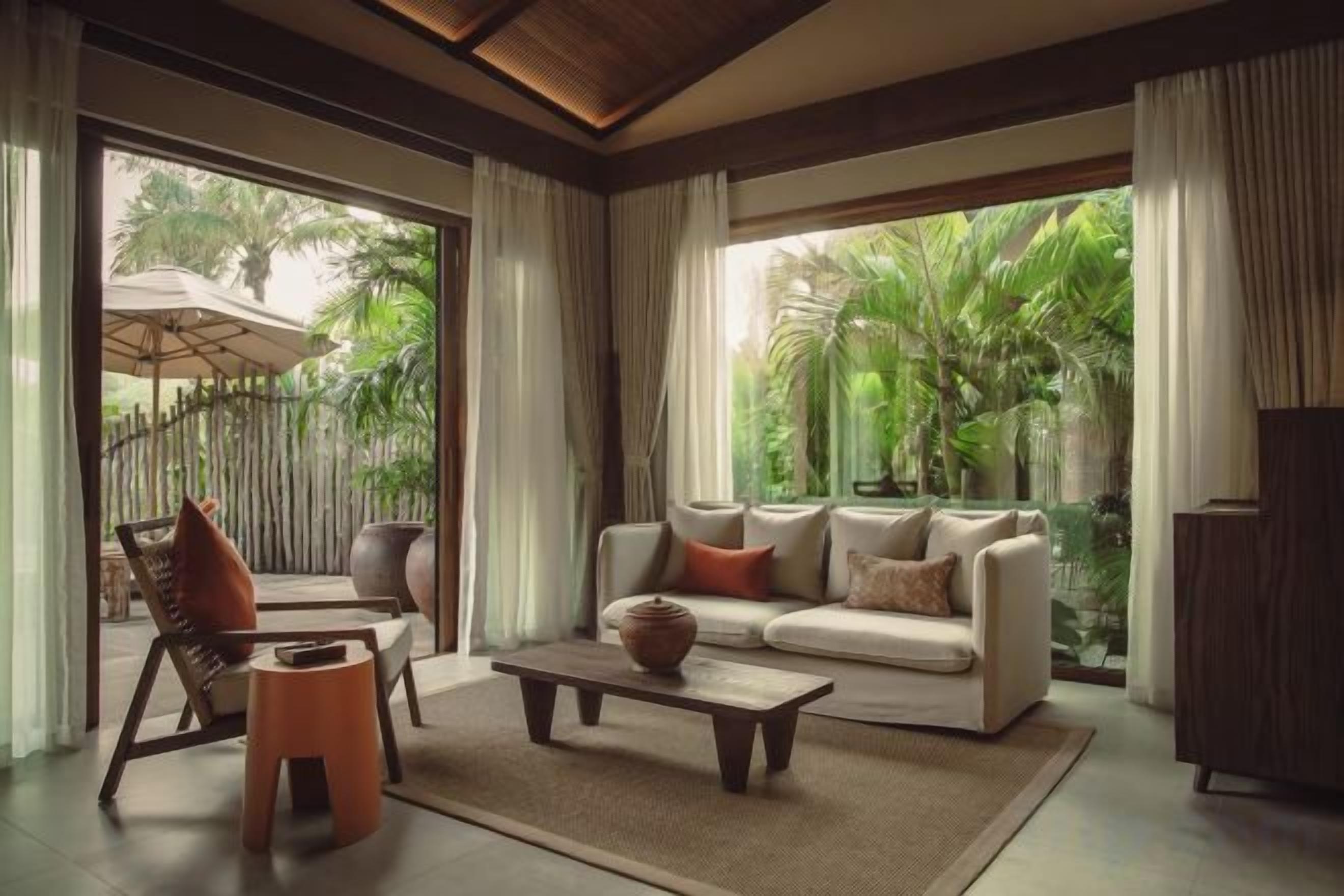 A modern living room with a sofa, coffee table, and chairs, large windows with a view of greenery.
