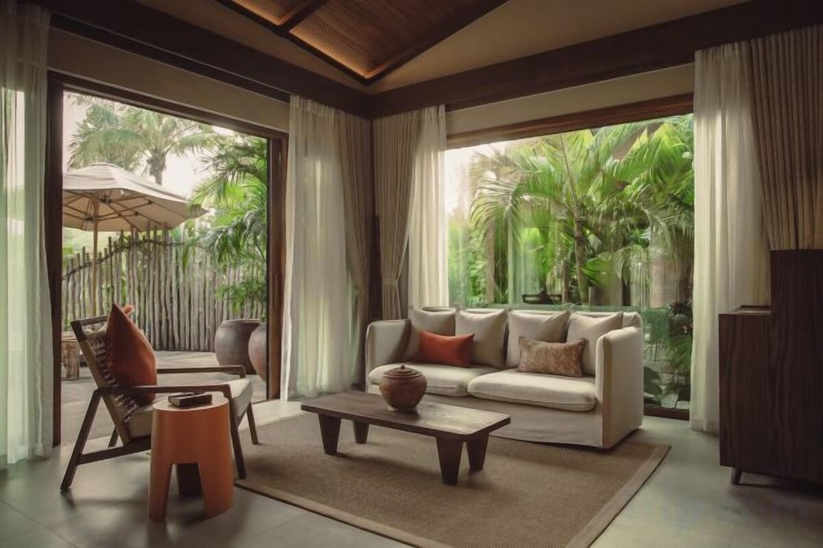 A modern living room with a sofa, coffee table, and chairs, large windows with a view of greenery.