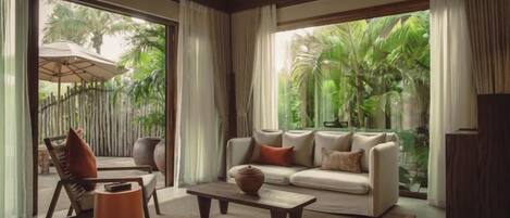 A modern living room with a sofa, coffee table, and chairs, large windows with a view of greenery.