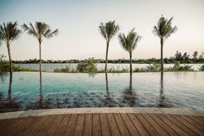 An infinity pool with a wooden deck, palm trees, and a view of the water. - Namia River Retreat - Wellness Inclusive Resort (Hoi An)
