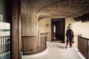 A curved wooden balcony with a railing, a person walking towards a door, and a view of the outside. - Namia River Retreat - Wellness Inclusive Resort (Hoi An)