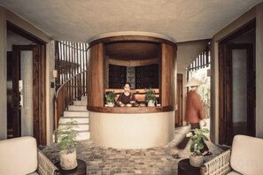 A reception area with a curved wooden desk, potted plants, and a staircase leading to upper floors. - Namia River Retreat - Wellness Inclusive Resort (Hoi An)